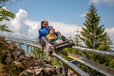 Vater und Sohn sitzen in einer Rodelbahn auf Schienen. | © JOSEF WIEGAND GMBH & CO.KG